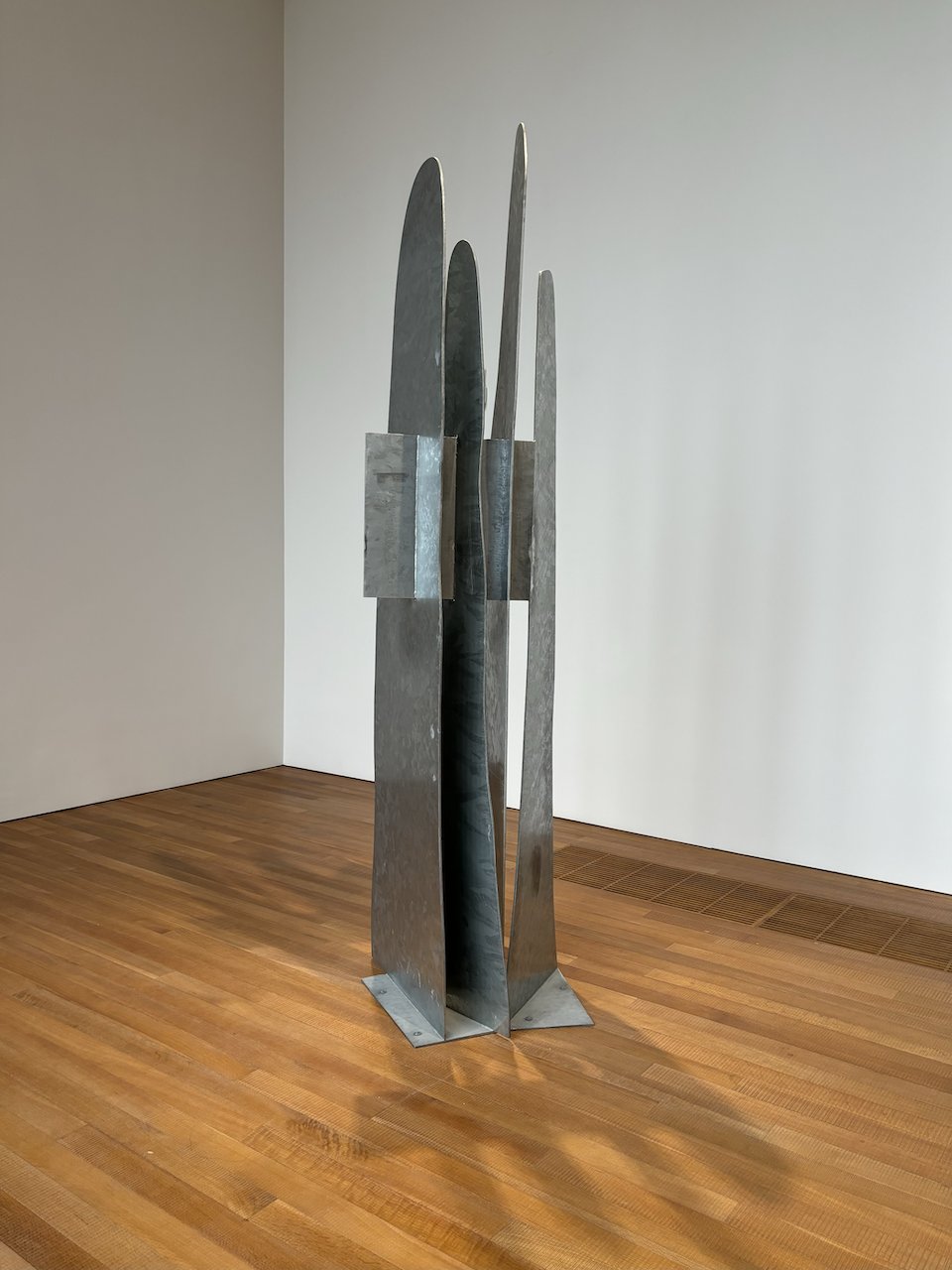A sculpture made of four long metal sheets in light grey resembles a waterfall cascading down a hill. A rectangular panel cuts across the four metal sheets on the upper half. The sculpture stands upright with the support of another rectangular panel on the floor.